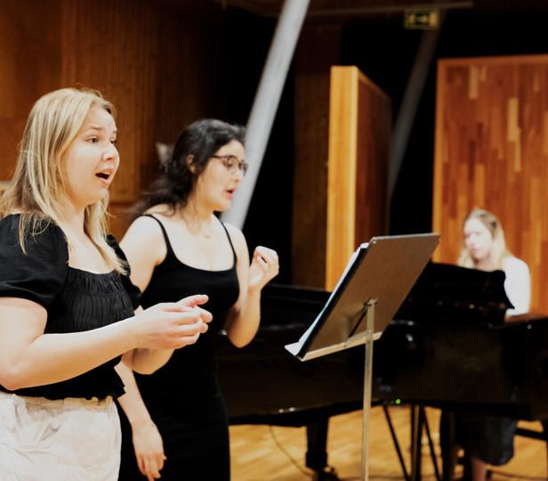 Two singers in a wood-paneled rehearsal studio, with a collaborative pianist at the keyboard, in the background. Both singers are focused on sheet music on a stand in front of them